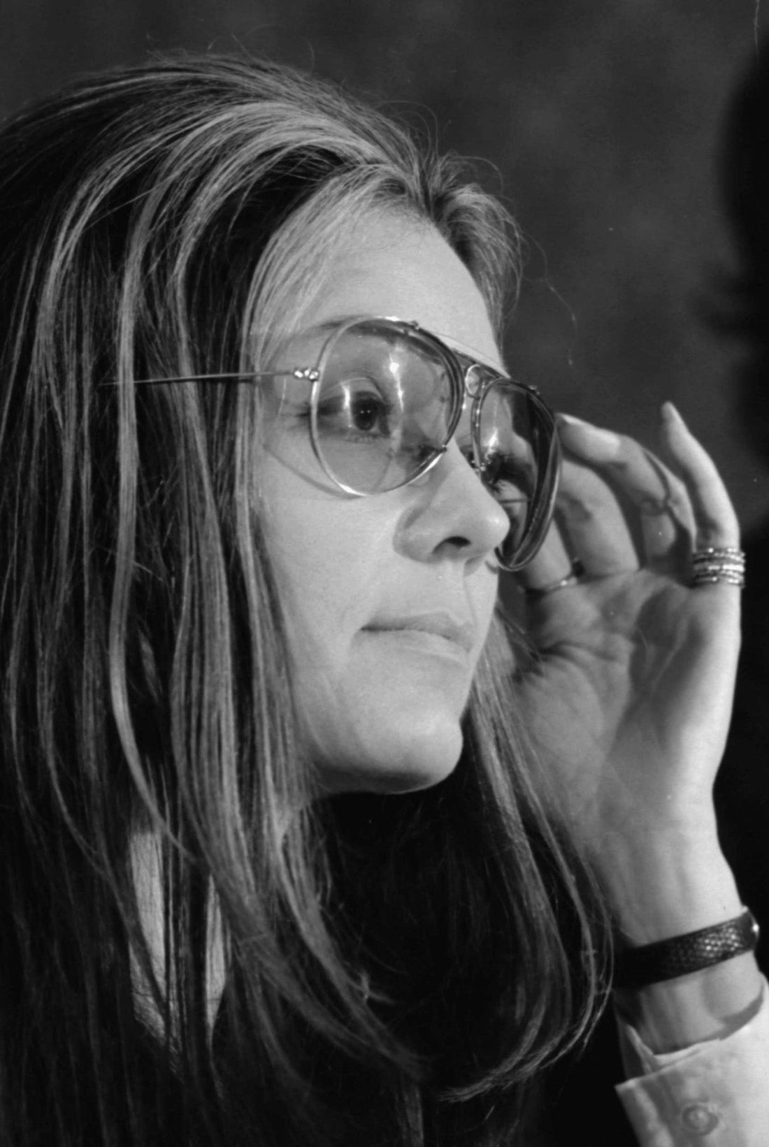 Black-and-white portrait of Gloria Steinem wearing large glasses and raising a hand to the frame.