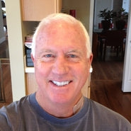 A close-up selfie of a smiling man with short white hair wearing a gray t-shirt, set against a home interior background with wooden floors and a doorway.